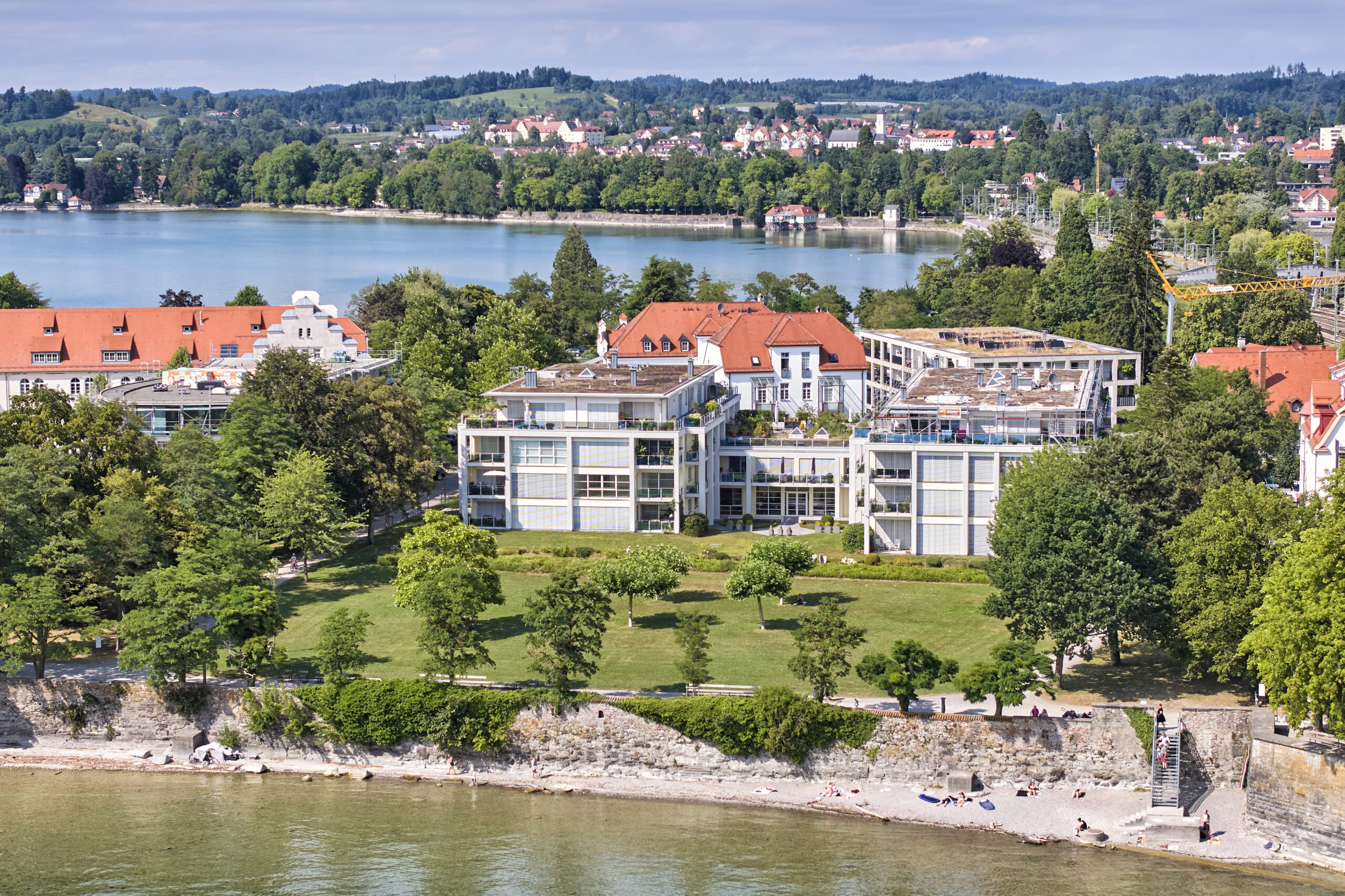 Luftaufnahme der Bodenseeklinik Lindau mit moderner Klinikgebäudefassade, angrenzender Parkanlage und Kiesstrand am Bodensee; im Hintergrund die Bucht mit Uferbebauung und bewaldeten Hügeln.
