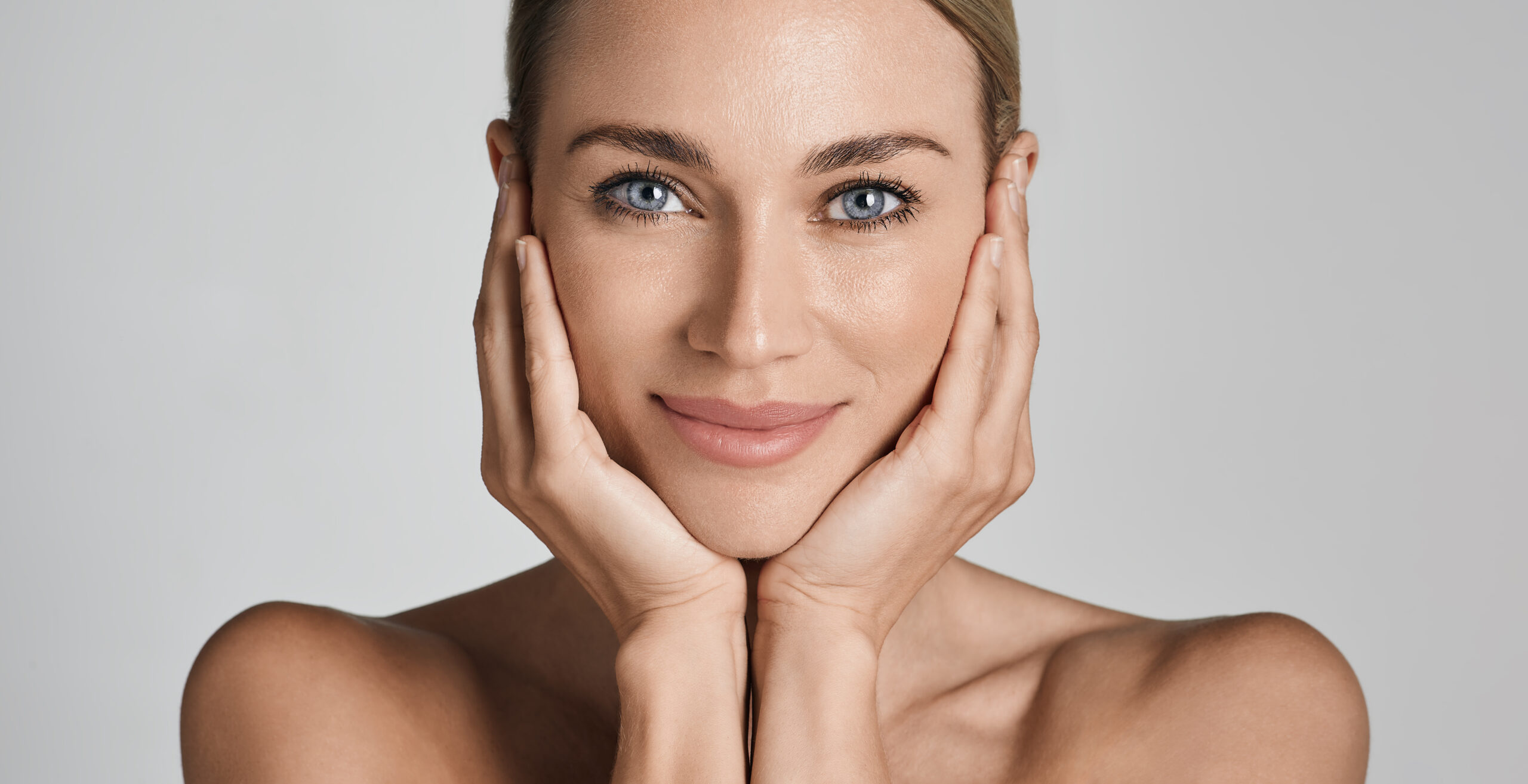 Skin care. Beautiful woman with healthy facial skin touching hands moisturized face skin, on light grey background. High quality Lächelnde blonde Frau mit strahlender, reiner Haut umrahmt ihr Gesicht mit beiden Händen.